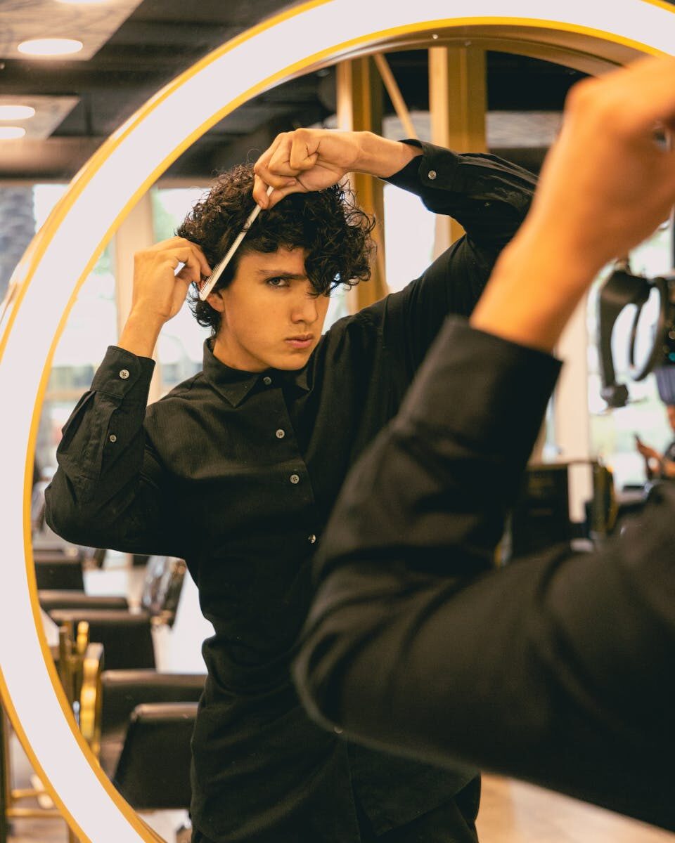 An adult man combing his curly hair in a stylish hair salon, looking focused in the mirror.