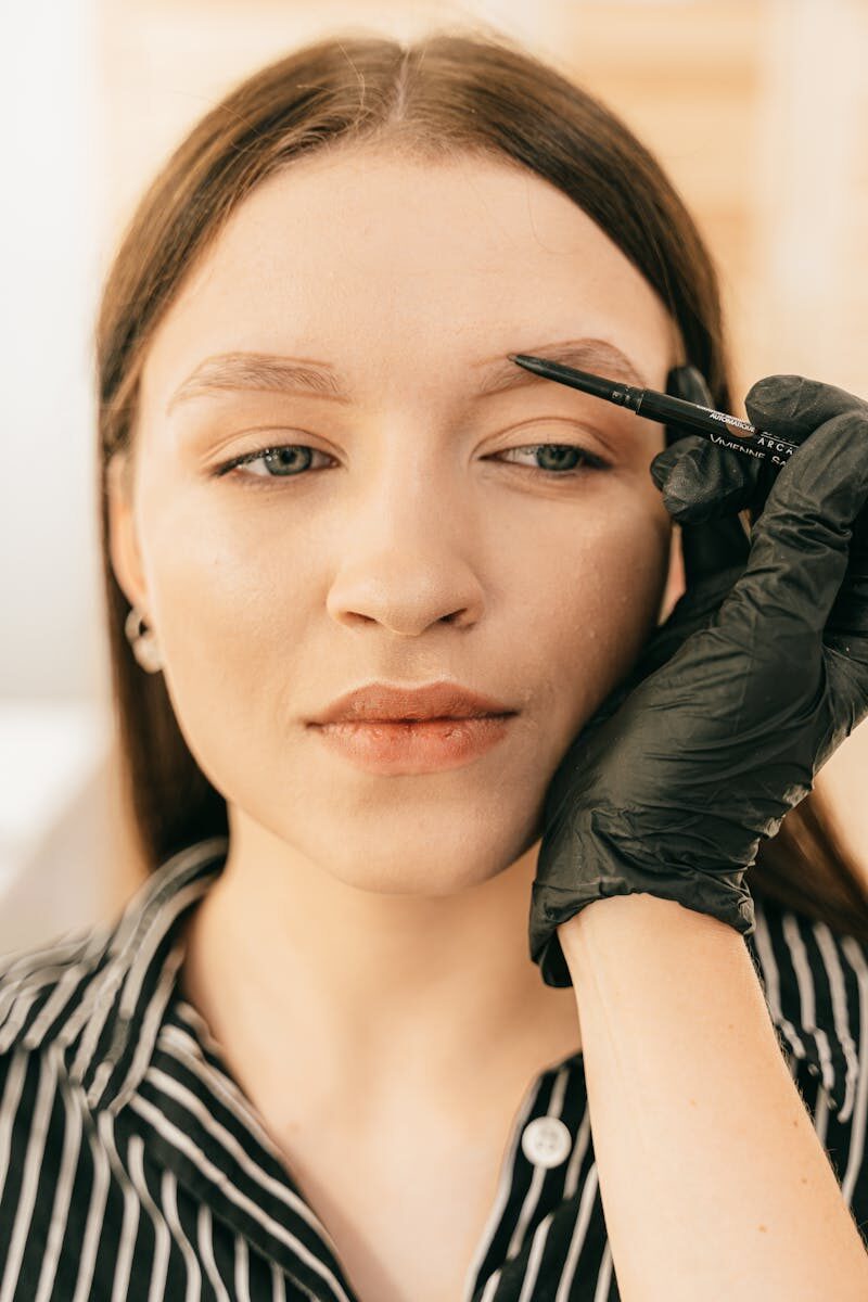 A beauty professional applies eyebrow makeup to a young woman indoors.